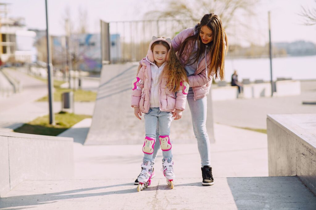 Full body cheerful young woman supporting daughter wearing protection pads while roller skating on concrete ramp during