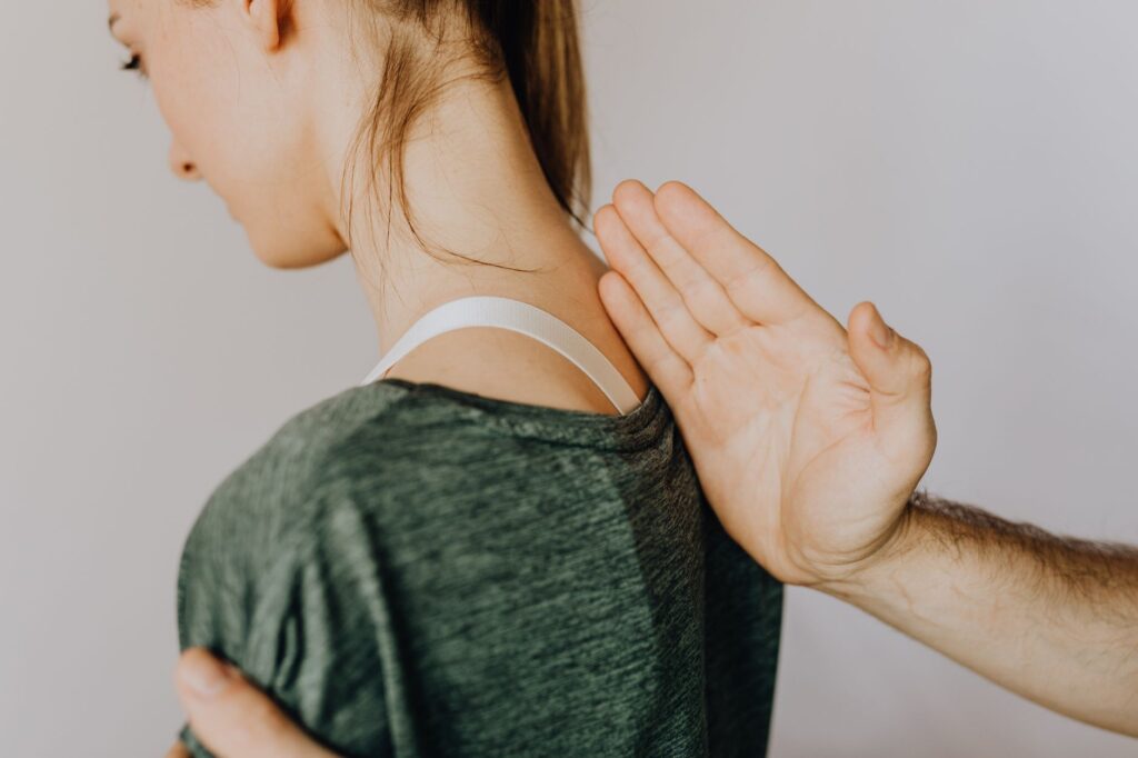 Side view of crop anonymous osteopath examining spine of slim young lady in casual wear near white wall in doctor office